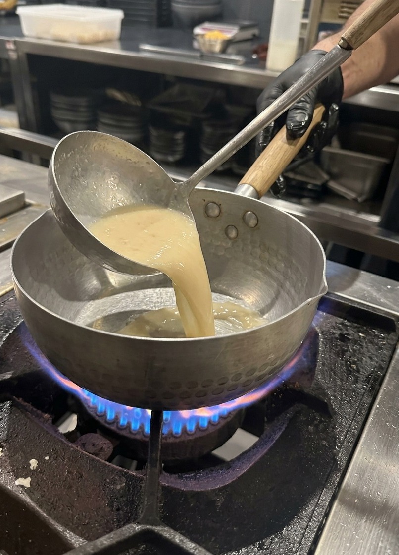 Une louche en métal versant un bouillon de porc clair dans une casserole sur une cuisinière à gaz.