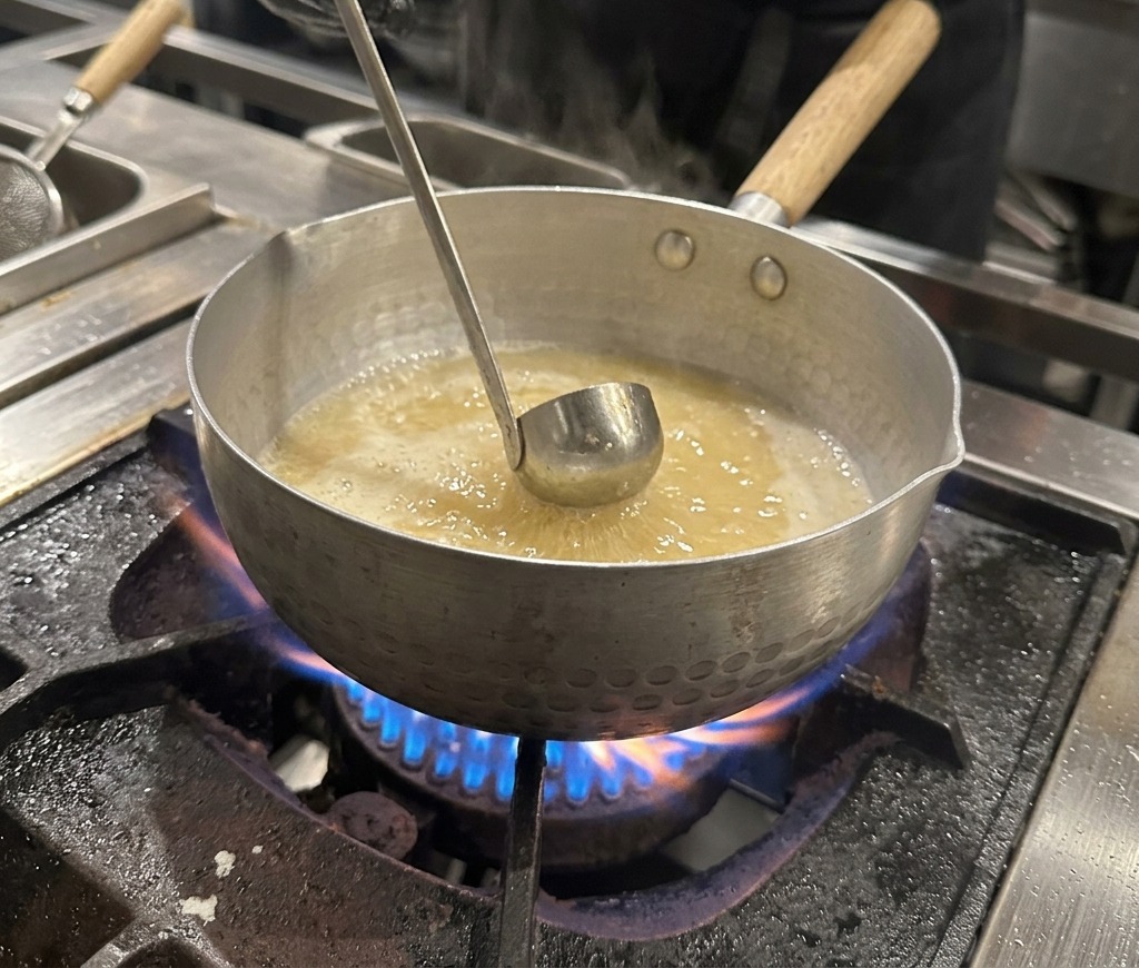 Une main gantée tenant un petit bol en métal, ajoutant de la sauce soja foncée dans un bouillon bouillonnant.