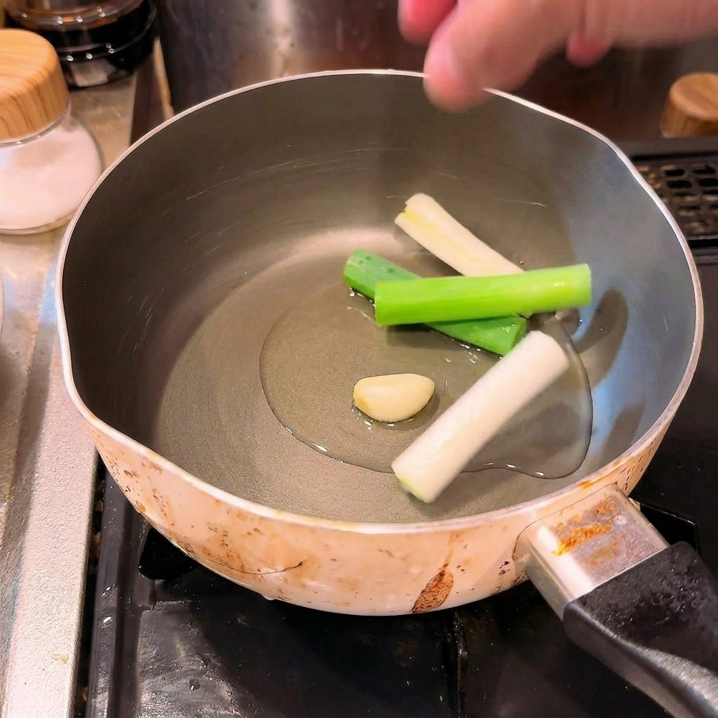 Une main déposant des morceaux doignons verts et une gousse dail dans une casserole dhuile froide.