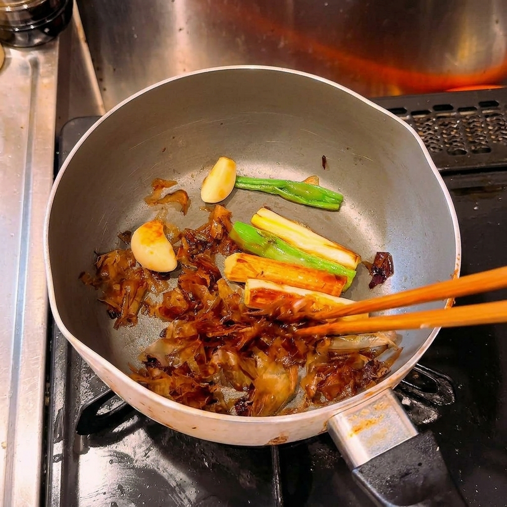 Des baguettes remuant des oignons verts, de lail et de la bonite intensément caramélisés et brunis dans une casserole.