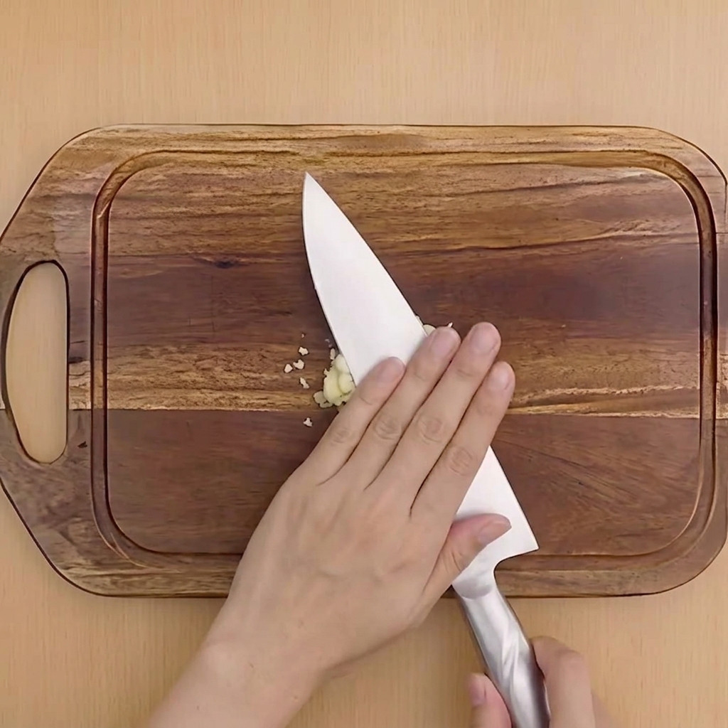 Une main pressant le plat dun couteau de chef pour écraser une gousse dail sur une planche en bois.