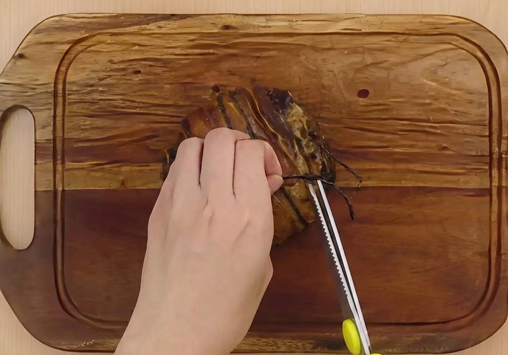 Des mains utilisant des ciseaux de cuisine pour couper la ficelle de boucher dun porc braisé sur une planche en bois.