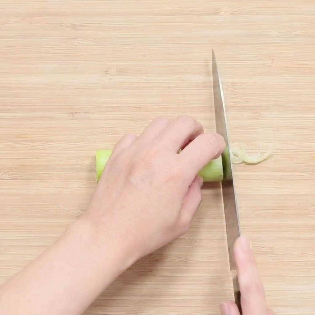 Des mains utilisant un couteau pour émincer finement la partie blanche dun oignon vert sur une planche.