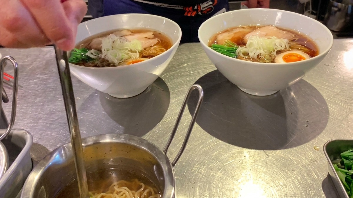 Une petite louche versant une huile aromatique claire sur un bol de ramen terminé.