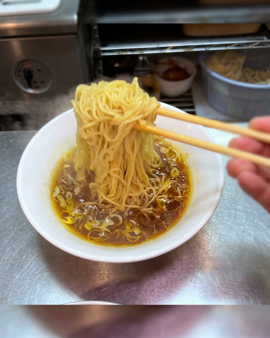 Baguettes soulevant une portion de nouilles ramen au-dessus dun bol de bouillon shoyu sombre.