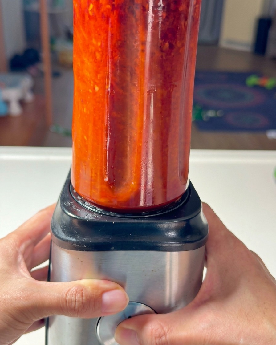 Deux mains fixant un bol de mixeur rempli de pâte de piment rouge sur sa base.