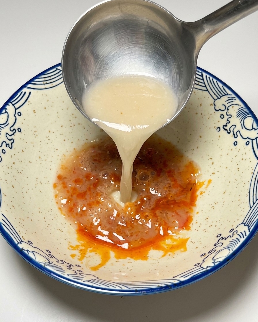 Bouillon Tonkotsu blanc et crémeux versé dune louche sur la pâte de piment dans un bol.