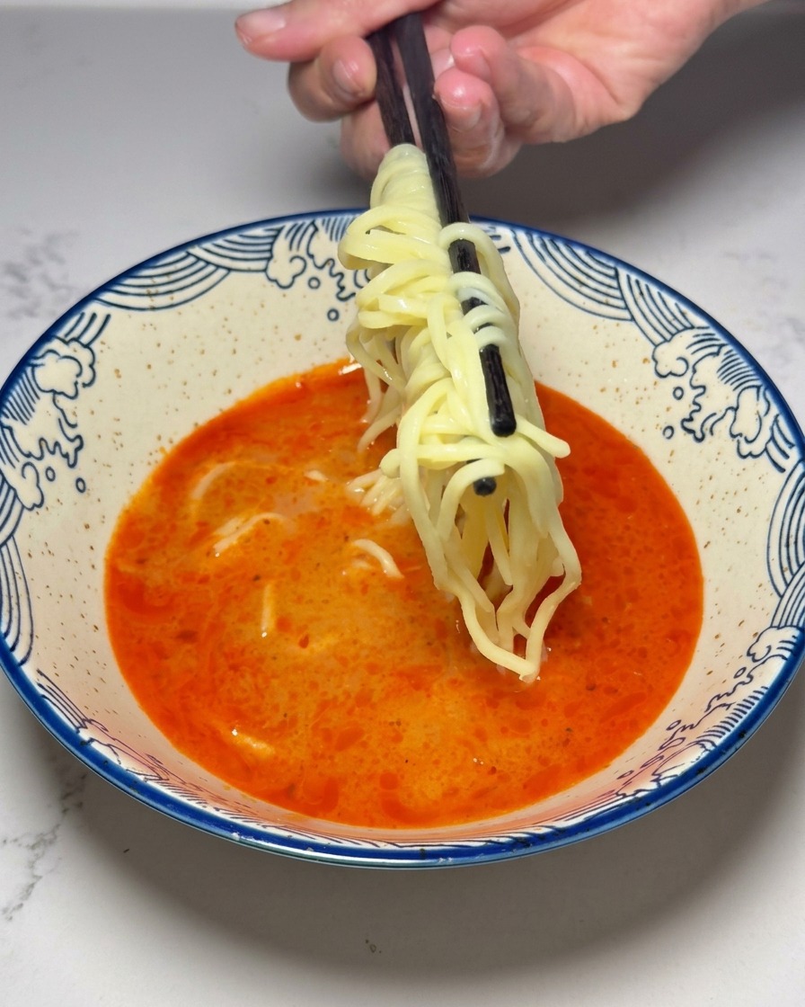 Nouilles ramen jaunes cuites plongées dans un bouillon rouge épicé avec des baguettes.