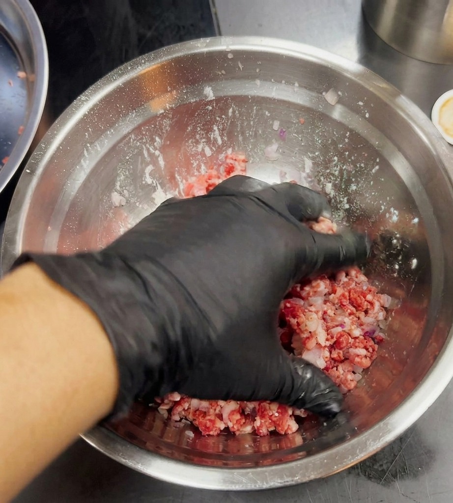 Une main gantée mélangeant doucement la viande hachée et les oignons mauves dans un bol argenté.