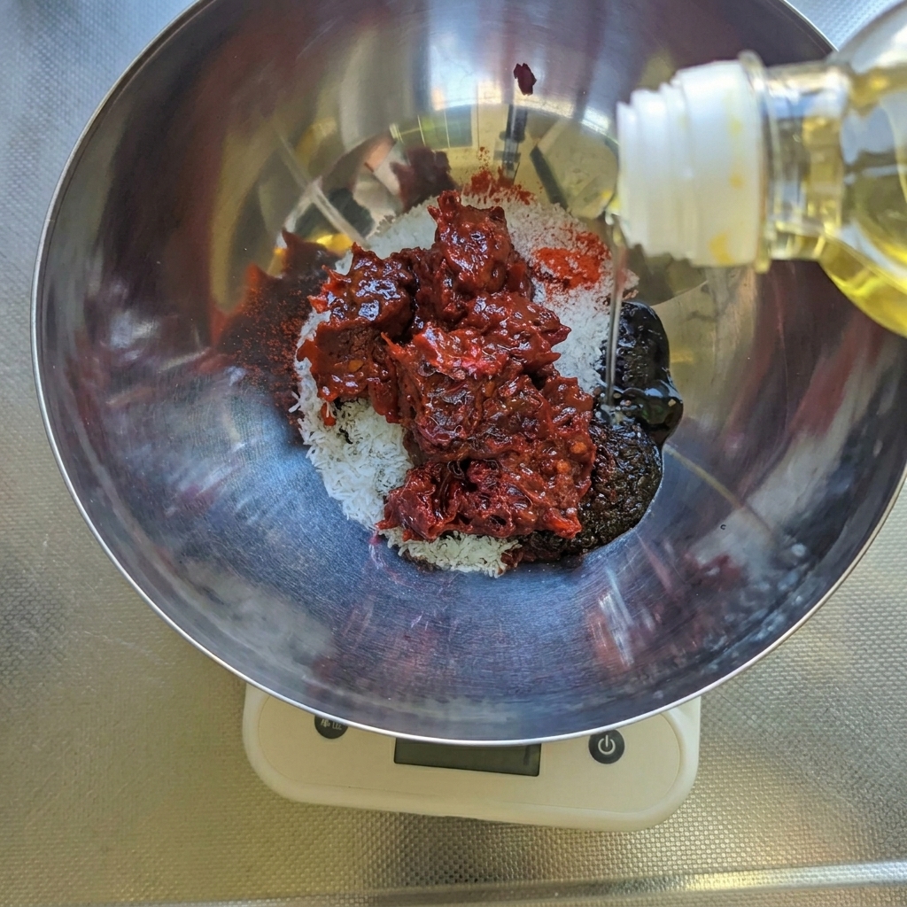 Huile végétale versée dans un bol contenant de la pâte de piment rouge, du sucre et des épices sur une balance.