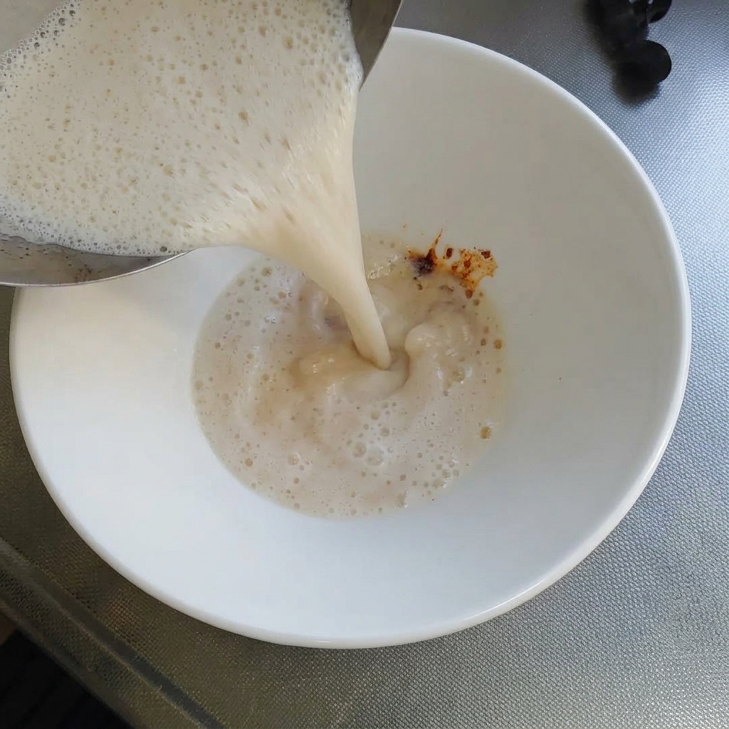Bouillon tonkotsu blanc et crémeux versé dune casserole dans un bol blanc contenant la pâte de piment rouge.