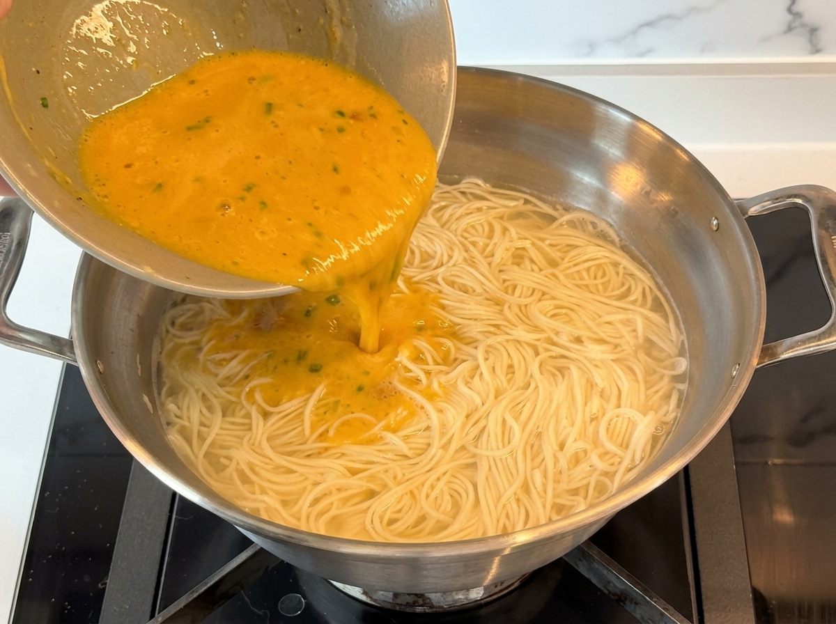 Une sauce crémeuse dorée versée dun petit bol dans une grande casserole remplie de nouilles et deau chaude.