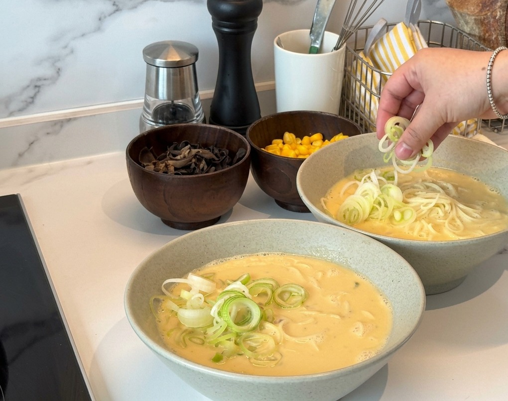 Une main saupoudrant des oignons verts ciselés sur deux bols de ramen au bouillon crémeux.