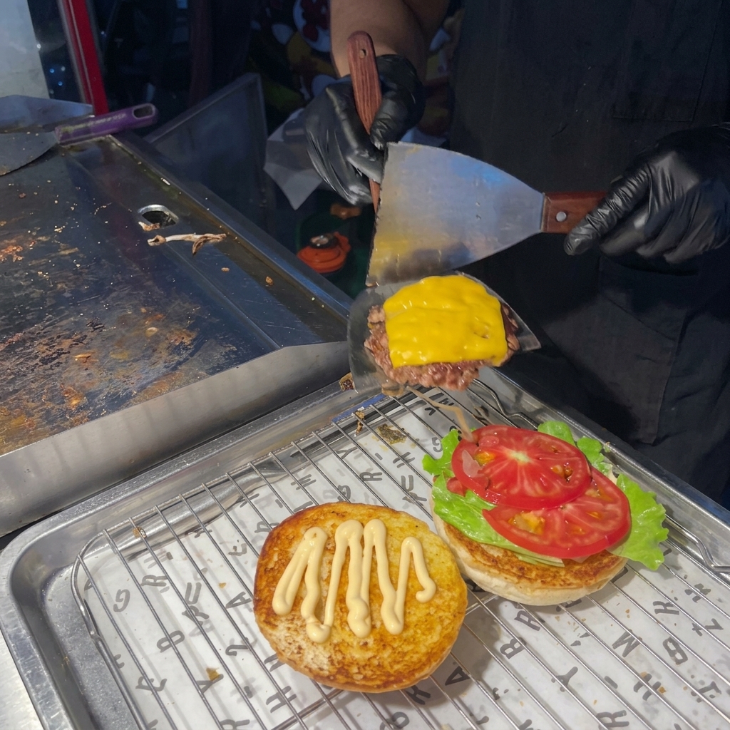 Une personne utilisant une spatule pour poser un steak au fromage fondu sur un burger garni de salade et tomate.