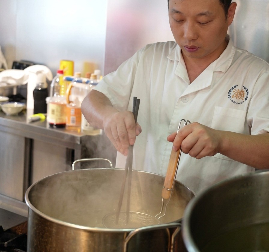 Un chef utilise de longues baguettes en bois pour remuer des nouilles ramen fraîches dans une passoire métallique plongée dans une marmite deau bouillante.