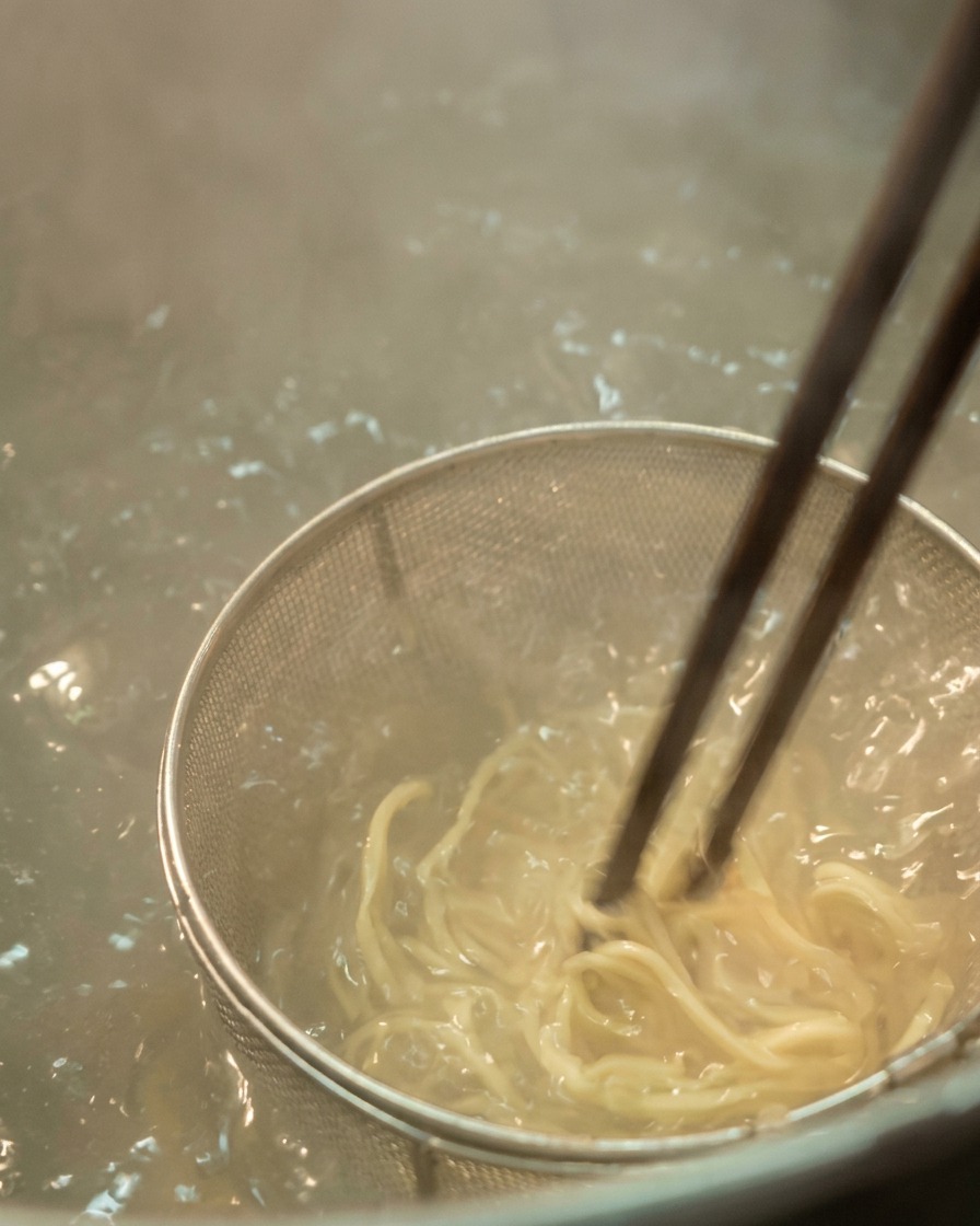 Gros plan sur des nouilles ramen claires remuées avec des baguettes sombres dans une passoire métallique immergée dans leau bouillante.