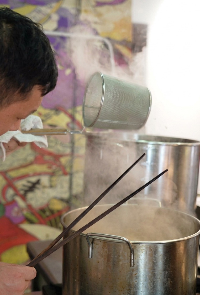 Un chef tenant une passoire à nouilles cylindrique au-dessus dune marmite fumante, la secouant pour égoutter les nouilles ramen.
