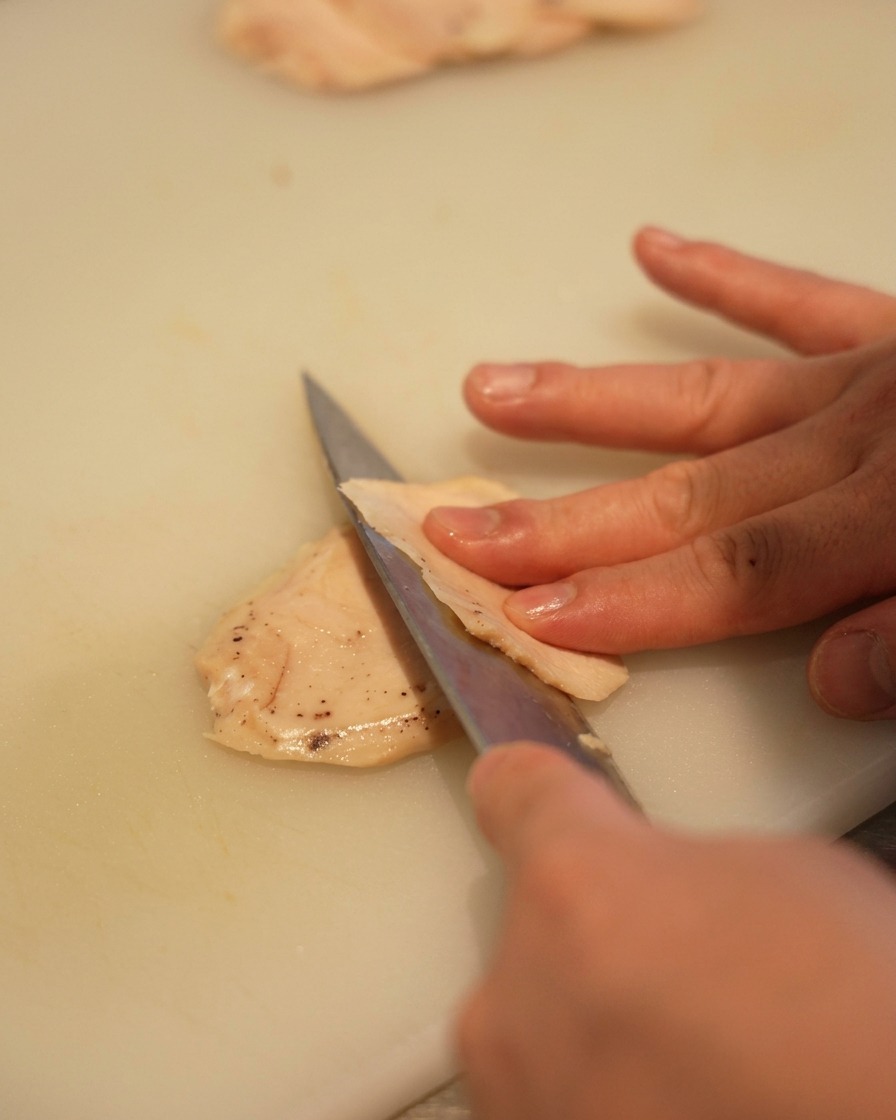 Gros plan dune main utilisant un couteau bien aiguisé pour couper un blanc de poulet pâle en fines tranches sur une planche à découper blanche.