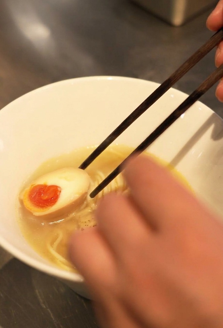 Gros plan de baguettes déposant un œuf mollet coupé en deux avec un jaune orange coulant dans un bol de bouillon et de nouilles.