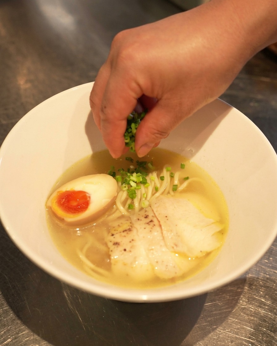 Une main saupoudrant des oignons verts fraîchement hachés sur un bol de ramen shio terminé, avec lœuf mollet et le poulet tranché.