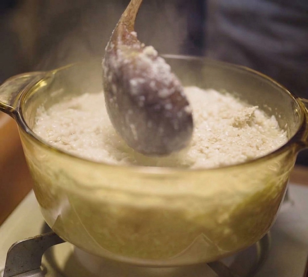 Une louche en bois remuant un bouillon blanc avec de fines particules de viande dans une marmite transparente.