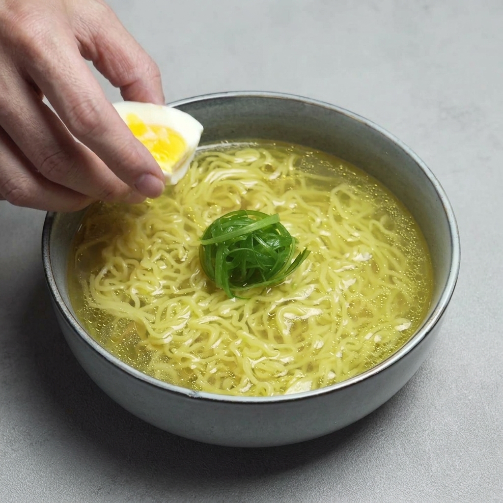 Main déposant une moitié dœuf mollet au jaune brillant dans le bol de ramen.