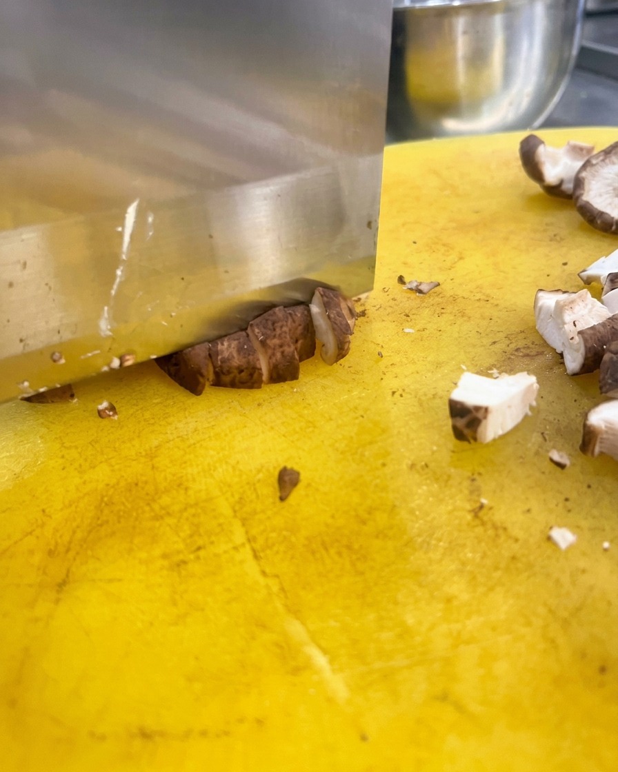 Un grand couteau de cuisine coupant des têtes de champignons shiitakés bruns en petits dés sur une planche jaune.