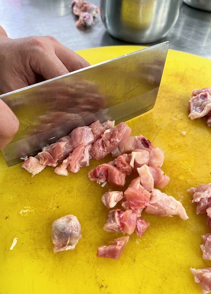 Une personne utilisant un couperet en métal pour couper du poulet cru en dés sur une planche en plastique jaune.