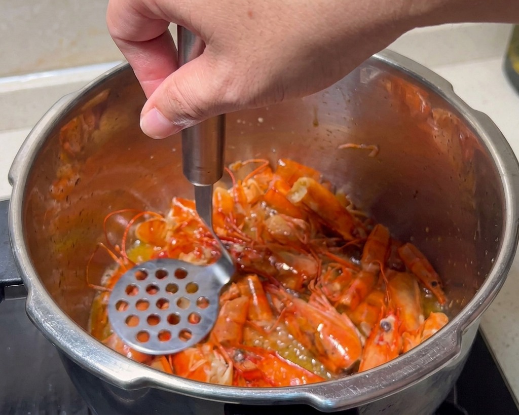 Un presse-purée métallique écrasant les têtes de crevettes rouges frites dans une marmite en inox.