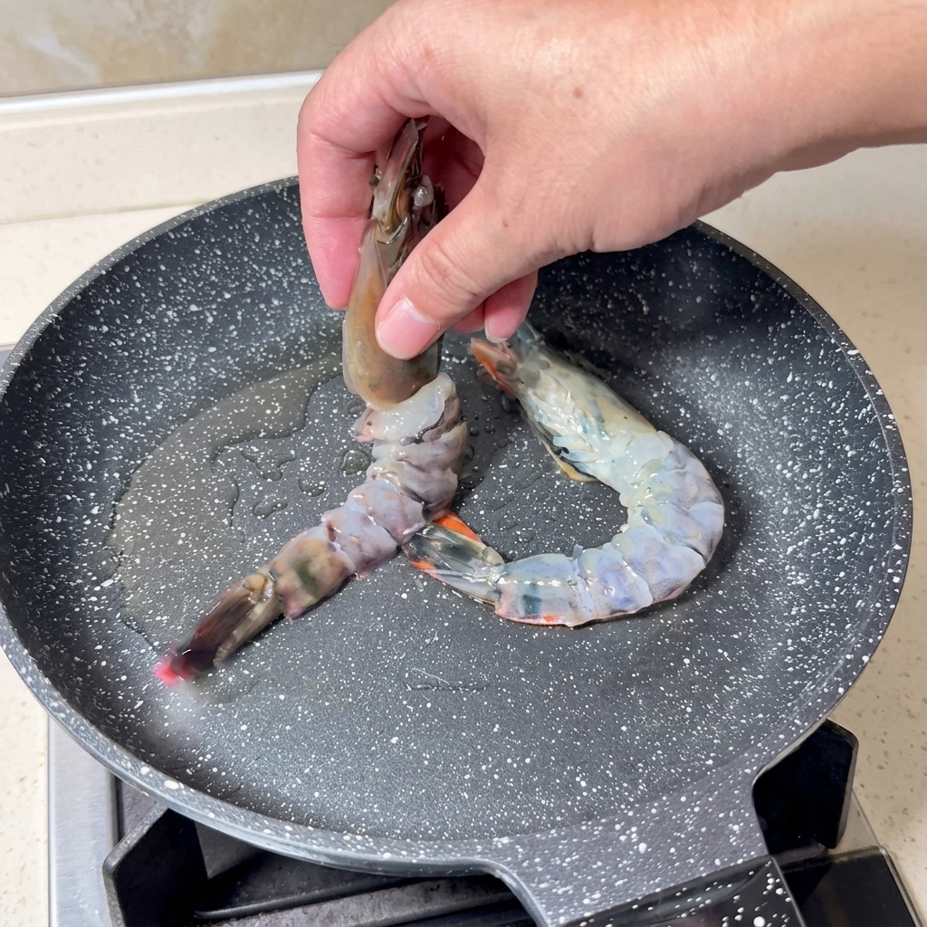 Main déposant de grosses crevettes crues en papillon dans une poêle chaude avec de lhuile.