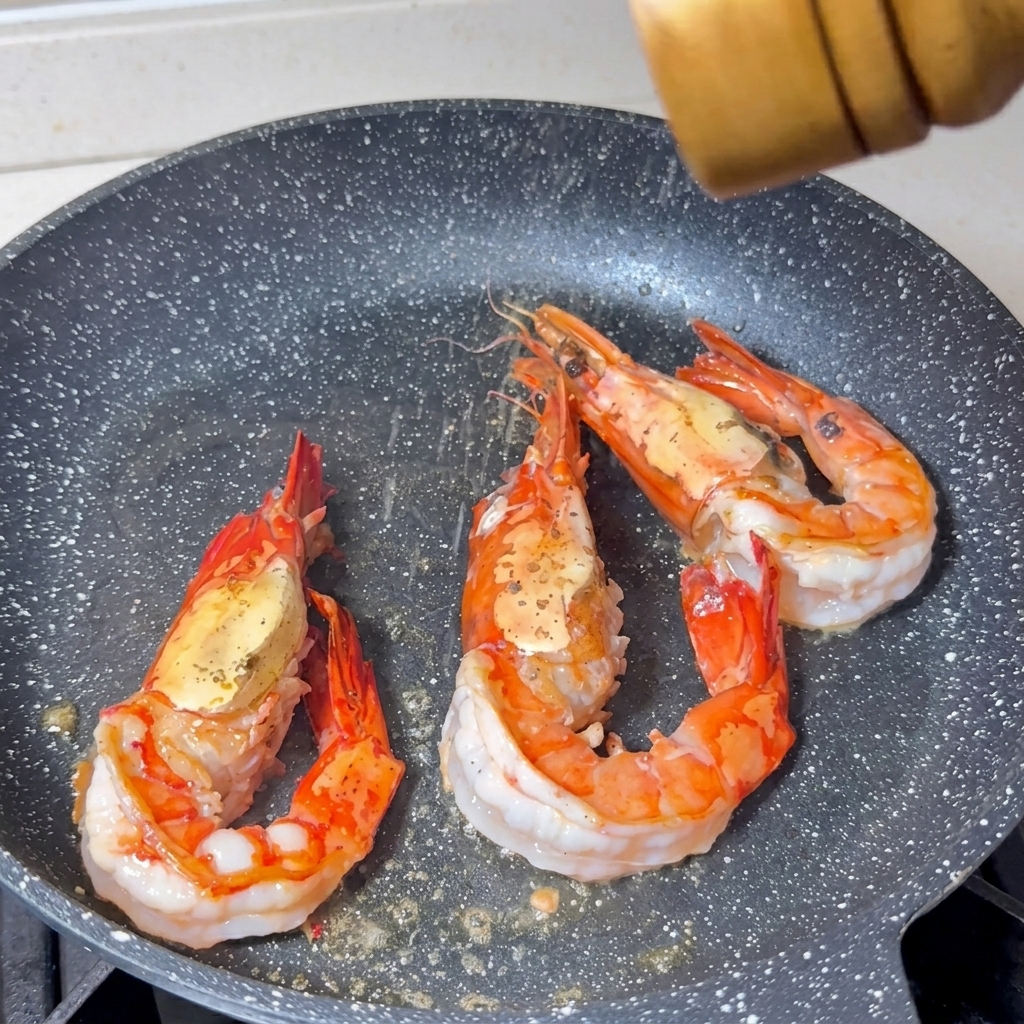 Sel et poivre moulus sur trois grosses crevettes en train de cuire dans une poêle.