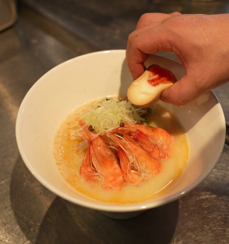 Une main déposant une moitié dœuf mollet brillant au jaune orangé fondant dans un bol de ramen déjà garni.
