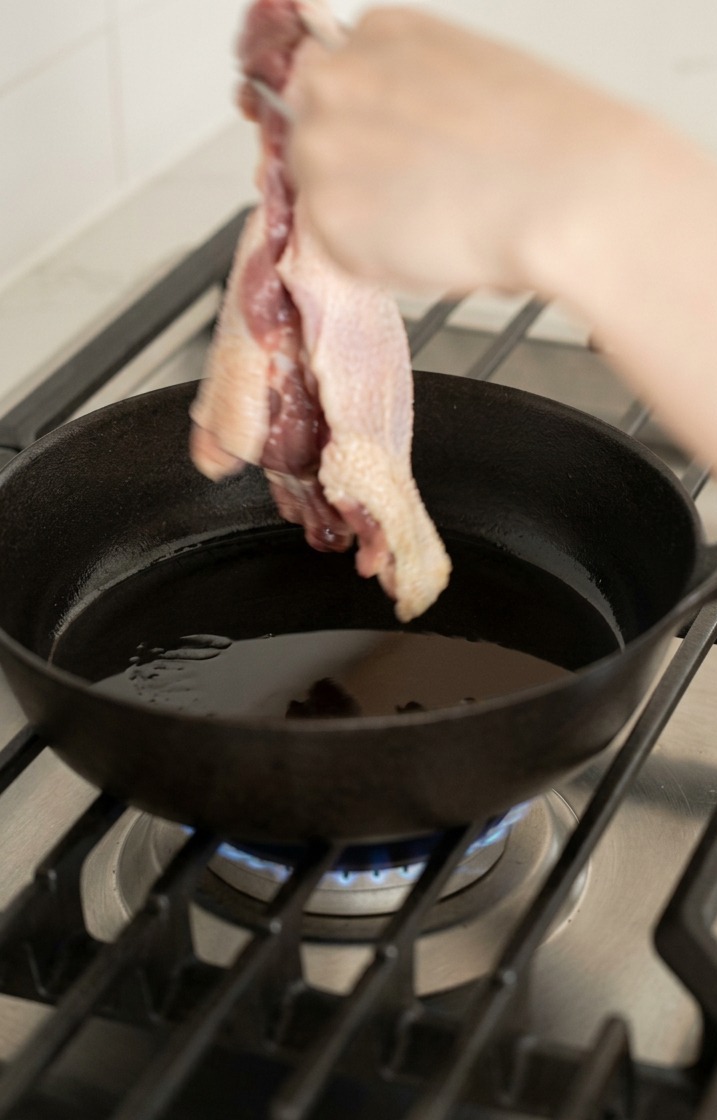 Une main utilisant des pinces pour placer une cuisse de canard crue côté peau dans une poêle en fonte chaude.