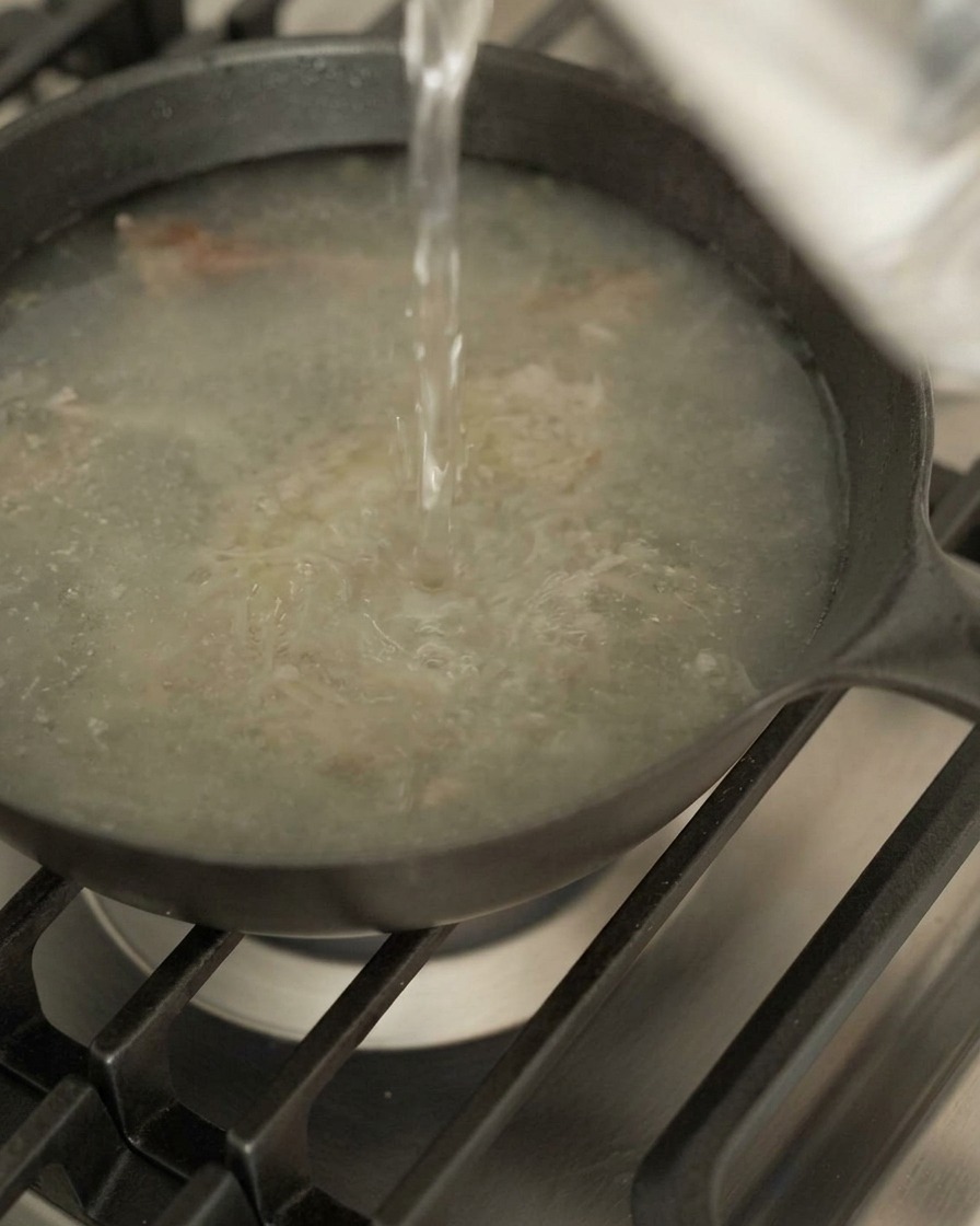 Eau versée dun récipient dans une poêle sombre contenant des sucs de viande sur une cuisinière à gaz.