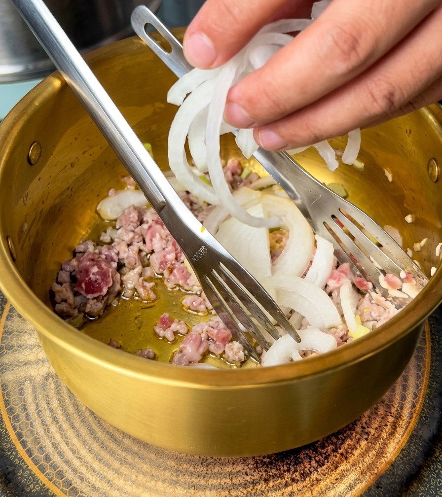 Une main ajoute des lamelles doignon blanc dans une marmite contenant le porc haché et les aromates.