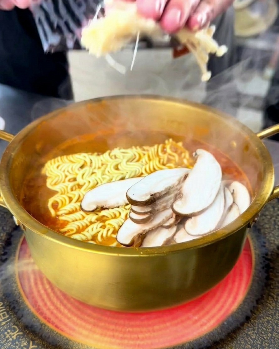 Une personne ajoute des enokis et des shiitakés émincés dans une marmite de ramen bouillante.