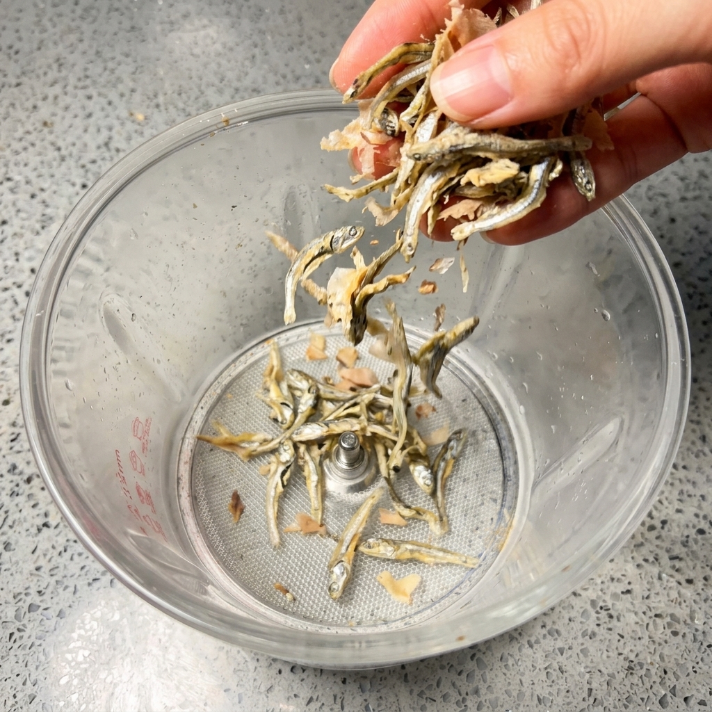 Une main déposant de petits anchois argentés séchés et des flocons de bonite rosâtres dans le bol dun mixeur.