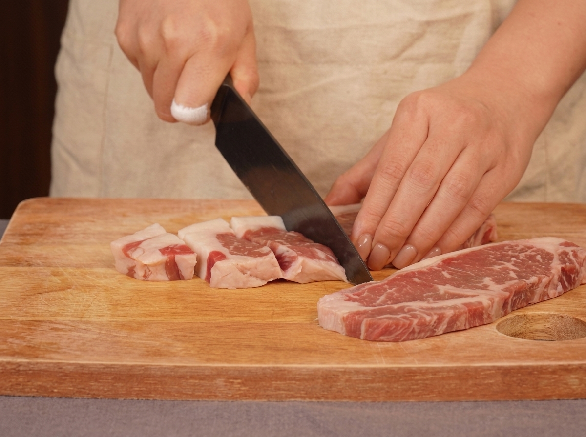 Une personne utilisant un couteau de chef pour couper du bœuf persillé cru en morceaux sur une planche en bois.