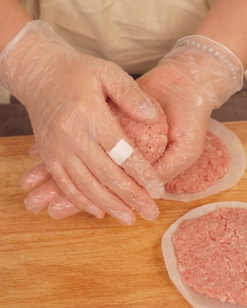 Une personne avec des gants en plastique lançant une boule de bœuf haché entre ses mains au-dessus dune planche à découper.