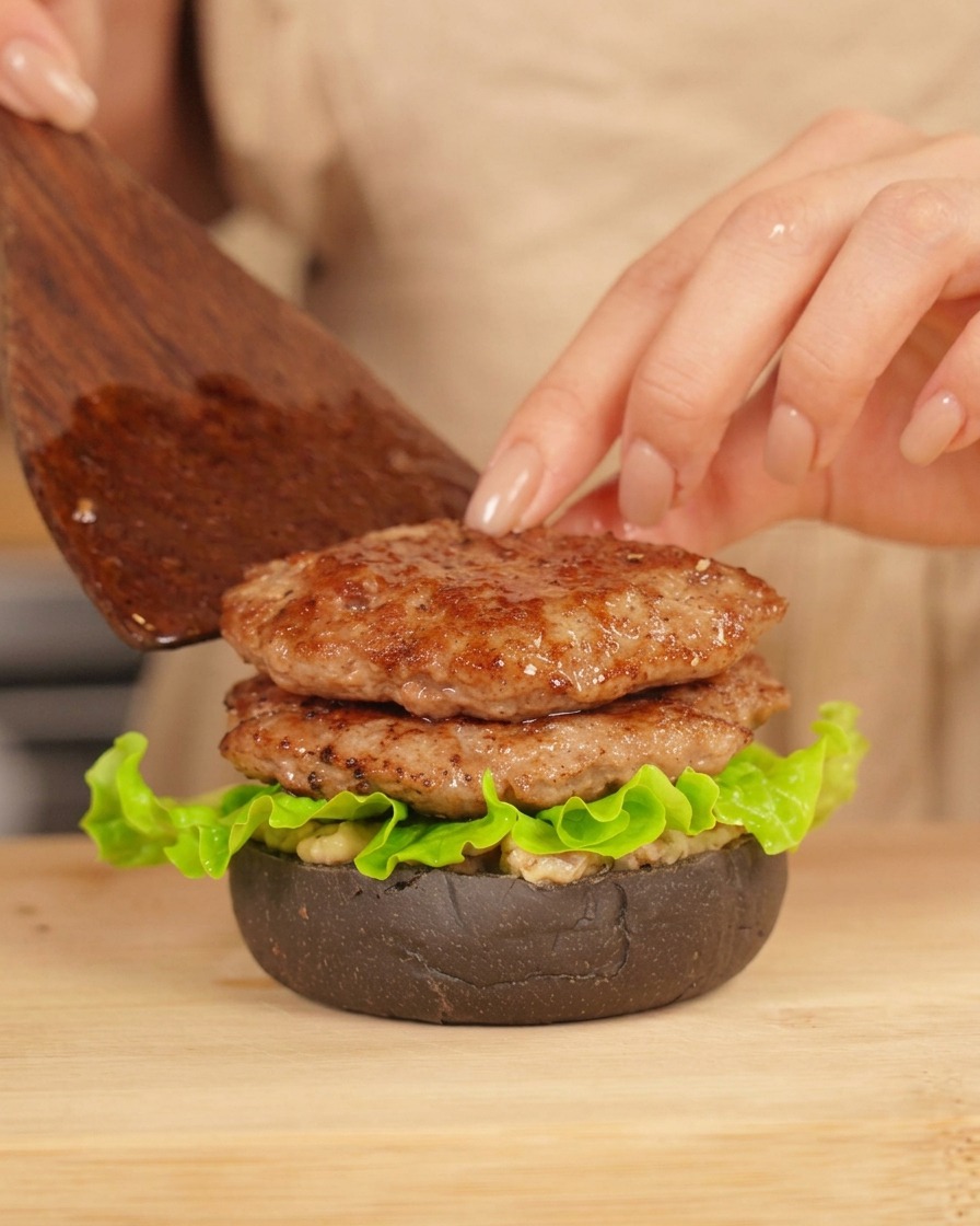 Une main utilisant une spatule en bois pour placer un deuxième steak grillé sur un burger avec pain noir et laitue.