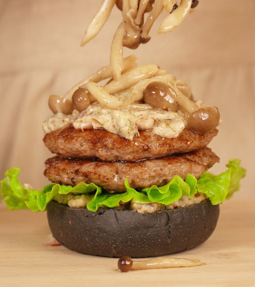 Une pince plaçant soigneusement des pleurotes sur un burger nappé de sauce crémeuse.