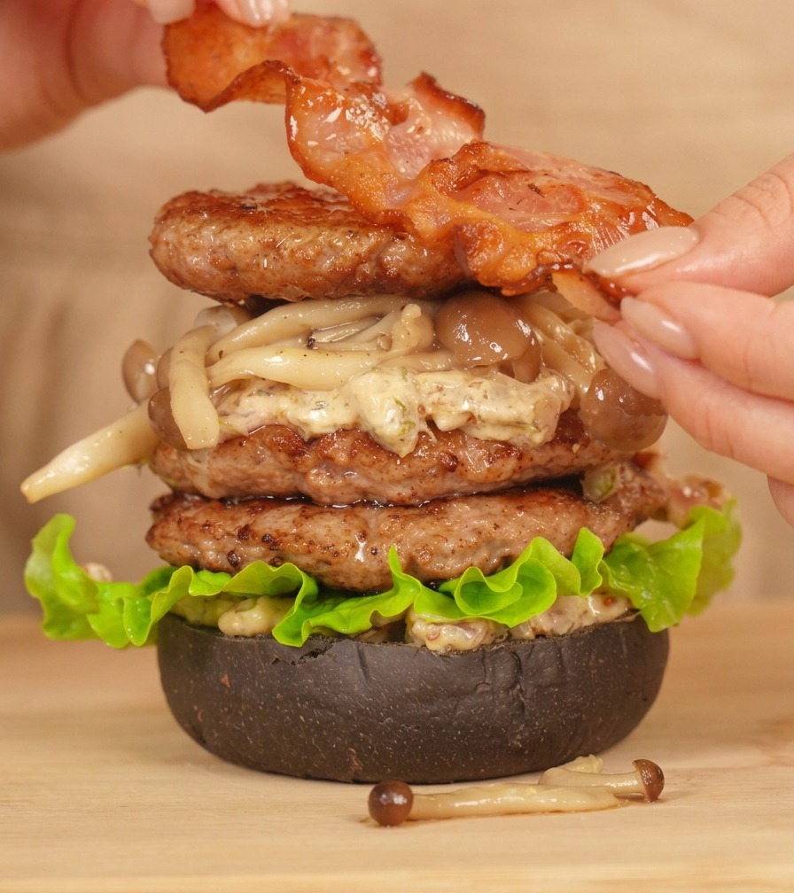 Mains plaçant une tranche de bacon parfaitement grillée au sommet dun grand double burger au bœuf.