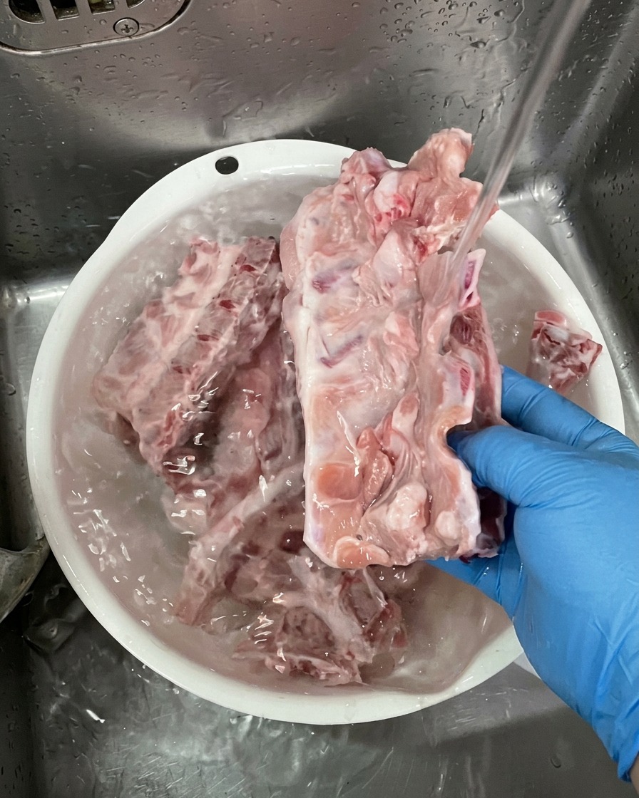 Une personne portant des gants bleus lave de grands os de porc crus sous un filet deau dans un bac en plastique blanc.