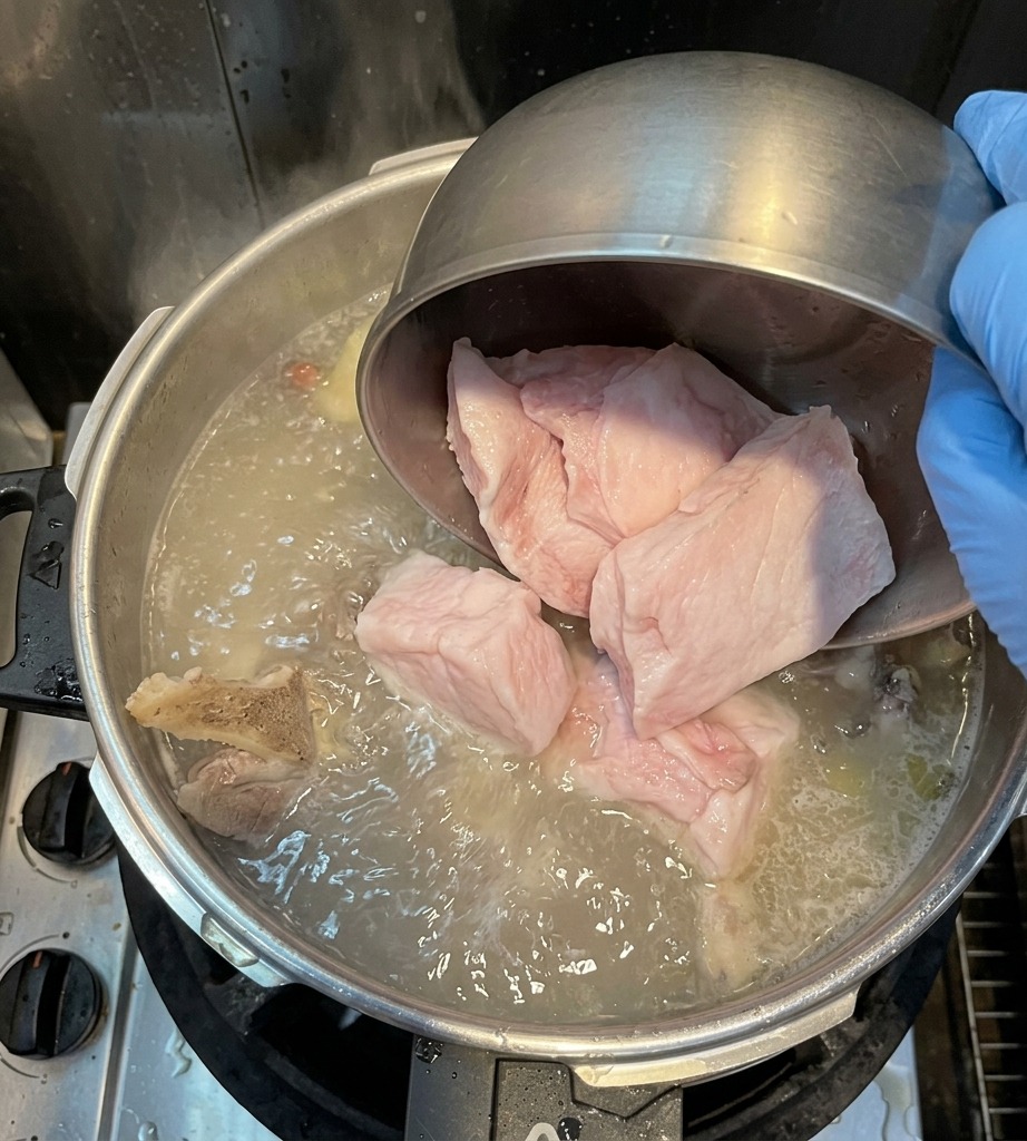 Une main gantée versant de gros morceaux de gras de dos de porc blanc depuis un bol en métal dans une marmite de bouillon en ébullition.