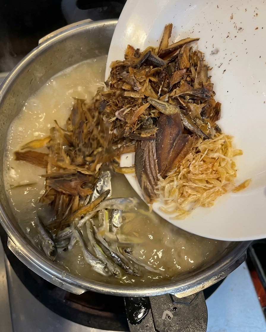 Des copeaux de poisson séché, de petites sardines argentées et de minuscules crevettes séchées versés depuis une assiette blanche dans un bouillon crémeux en ébullition.