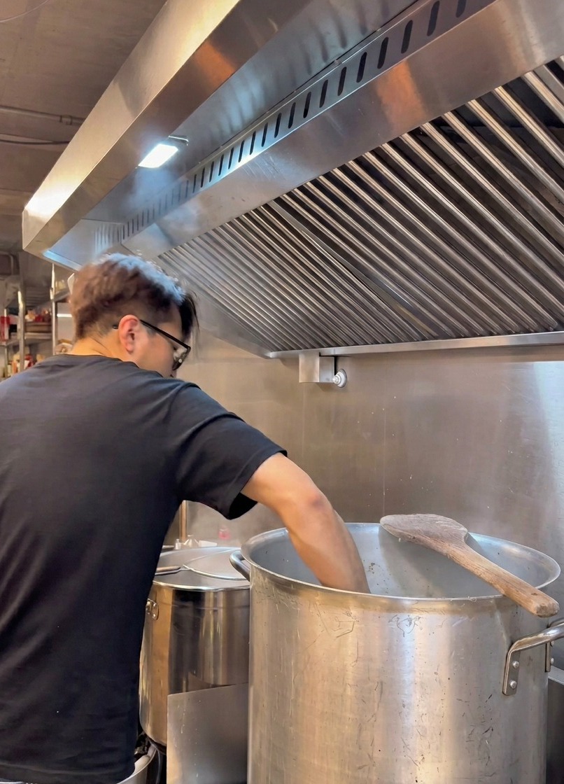 Un chef en t-shirt noir se penche sur une grande marmite en métal pour filtrer un bouillon blanc crémeux à travers une passoire métallique sous une hotte de cuisine industrielle.