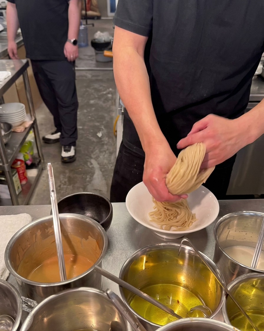 Un chef utilise ses deux mains pour plier et presser un nid de nouilles ramen épaisses et claires dans un bol en céramique blanche.