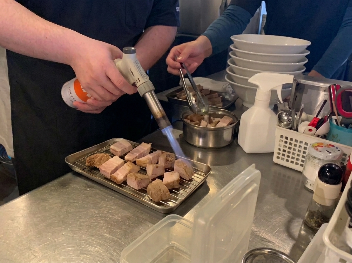 Un chef utilisant un chalumeau de cuisine pour caraméliser des tranches de poitrine de porc braisée sur une grille métallique.