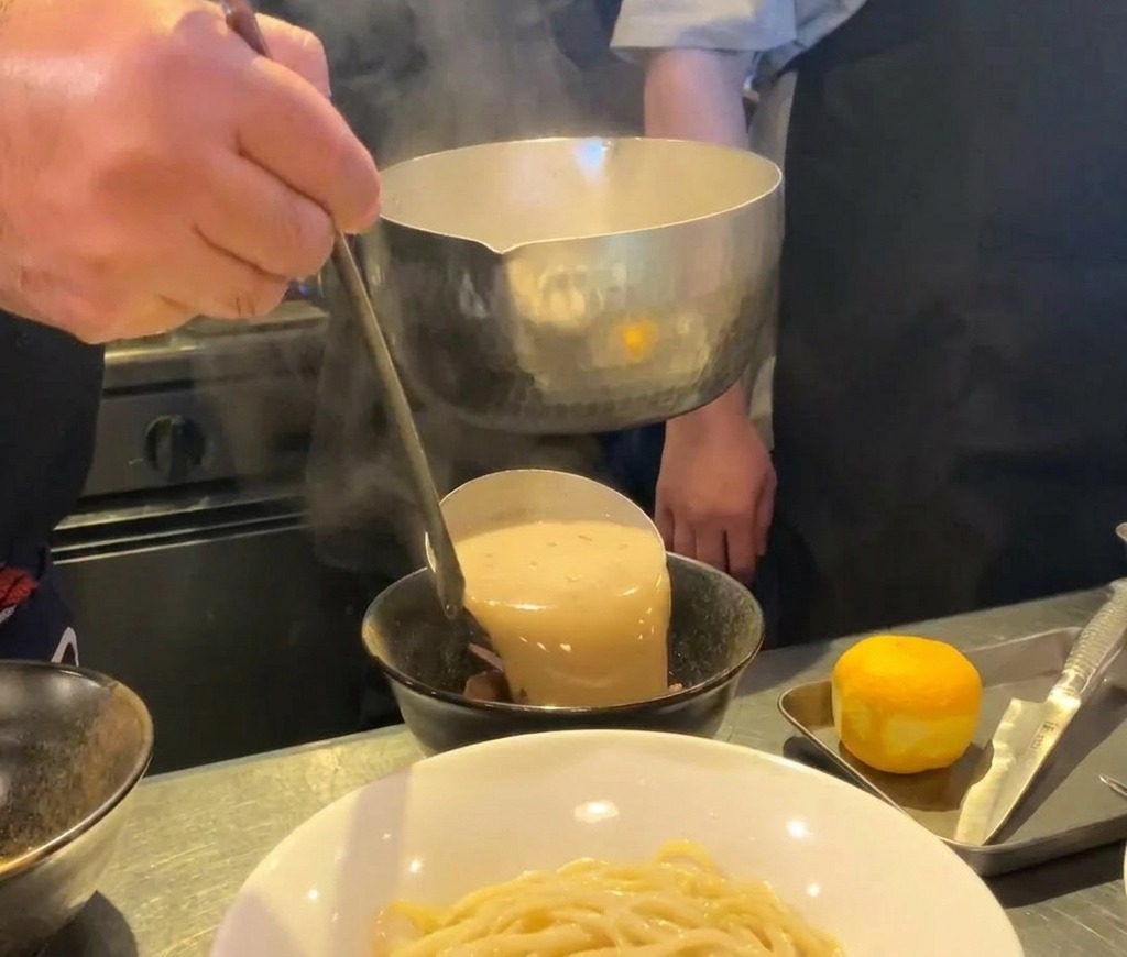 Bouillon de ramen aux fruits de mer épais et crémeux versé dune louche métallique dans un bol en céramique noire.