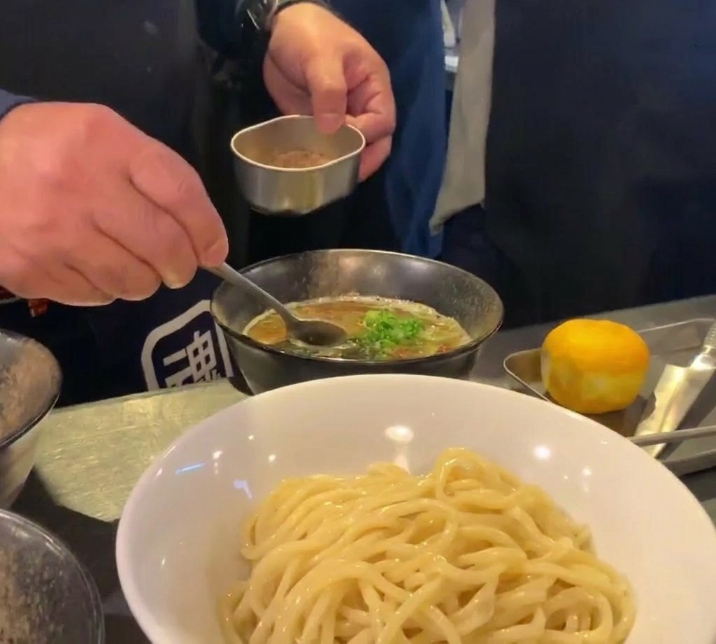 Un chef utilisant un petit récipient en métal et une cuillère pour saupoudrer une fine poudre de poisson brune sur un bol de soupe de trempage riche et sombre.